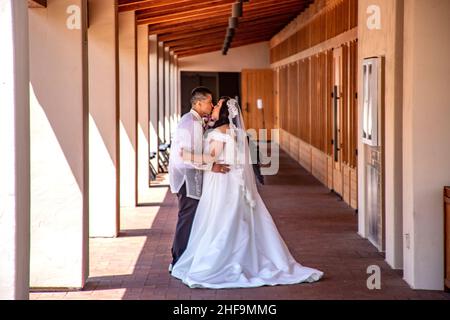 Nach der Hochzeitszeremonie genießen eine hispanische Braut und ein Bräutigam einen Moment Privatsphäre und teilen sich einen Kuss vor einer katholischen Kirche in Südkalifornien. Stockfoto