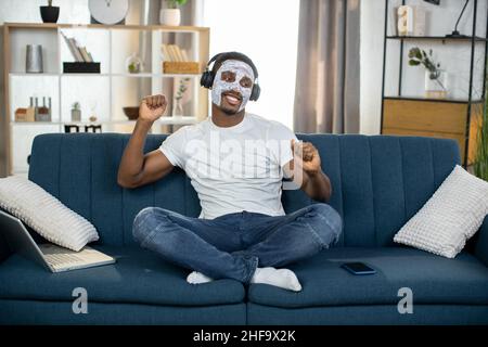 Entspannter afroamerikanischer Mann mit weißer Tonmaske, sitzt zu Hause auf dem Sofa und hört Musik in Kopfhörern, tanzt und genießt Lieder Stockfoto