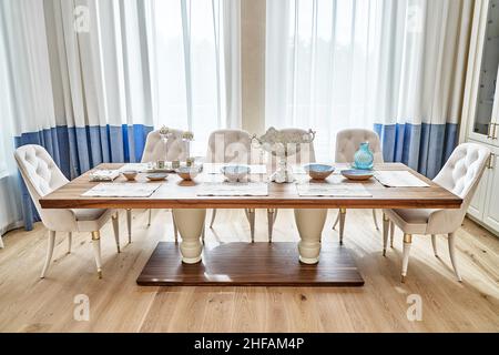 Luxuriöser Vintage-Esstisch aus massivem Nussbaum- und Furnierholz mit festlichem Ambiente und Sesseln in der Nähe des Fensters in einem geräumigen Zimmer Stockfoto