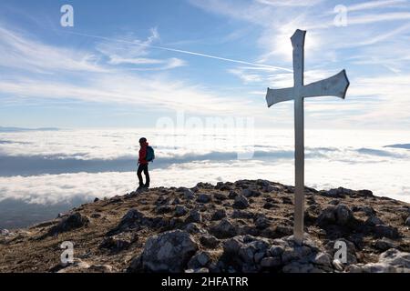 Frau, die am Kreuz auf dem Gipfel des Kucelj-Gebirges, über dem Nebel im Vipava-Tal, Slowenien, steht Stockfoto