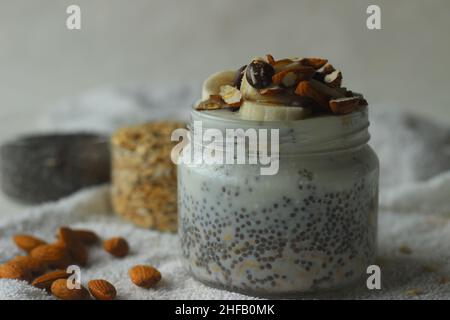 Hafer über Nacht. Frühstück im Glas. Hergestellt durch Einweichen von gerolltem Hafer und Chiasamen in Milch, serviert mit gehackter Banane, Mandeln, Choco-Chips und einem Nieselregen Stockfoto