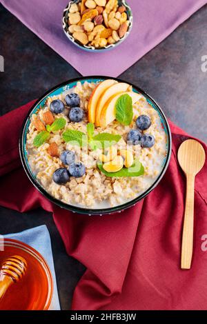 Haferbrei mit Joghurt, Beeren und Nüssen auf burgunderfarbener Serviette. Schüssel mit Haferflocken, Blaubeeren, Äpfeln und Honig auf dunklem Grund. Gesunde Brea Stockfoto