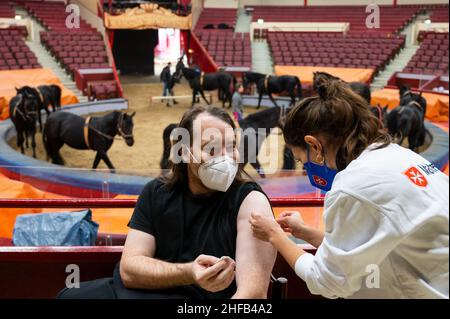 München, Deutschland. 15th Januar 2022. Die Vaccinologin Caroline Klima impft Thomas Frauendorfer im Circus Krone gegen das Corona-Virus, während Pferde im Ring im Hintergrund trainieren. Kredit: Lennart Preiss/dpa/Alamy Live Nachrichten Stockfoto