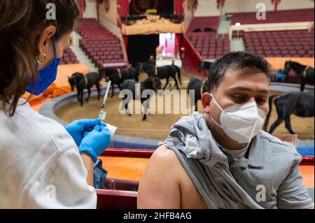 München, Deutschland. 15th Januar 2022. Die Vaccinologin Caroline Klima impft Deshad Rahim M. Rashid im Circus Krone gegen das Corona-Virus, während Pferde im Hintergrund im Ring trainieren. Kredit: Lennart Preiss/dpa/Alamy Live Nachrichten Stockfoto