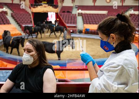 München, Deutschland. 15th Januar 2022. Die Vaccinologin Caroline Klima impft Thomas Frauendorfer im Circus Krone gegen das Corona-Virus, während Pferde im Ring im Hintergrund trainieren. Kredit: Lennart Preiss/dpa/Alamy Live Nachrichten Stockfoto