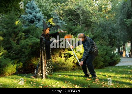 Ein junges, schönes Mädchen in Hexenkostüm versucht, einen Mann zu verzaubern, der einen Gartenbesen in den Händen hält. Holloween. Emotionskonzept. Stockfoto