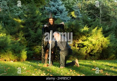 Ein junges, schönes Mädchen in Hexenkostüm versucht, einen Mann zu verzaubern, der einen Gartenbesen in den Händen hält. Holloween. Emotionskonzept. Stockfoto