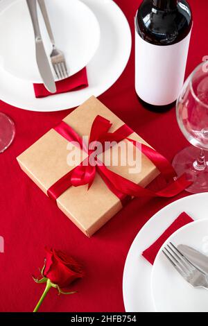 Romantisches Abendessen am Valentinstag. Geschenkbox, Teller mit Besteck, Flasche Wein, Gläser auf rotem Tisch Stockfoto