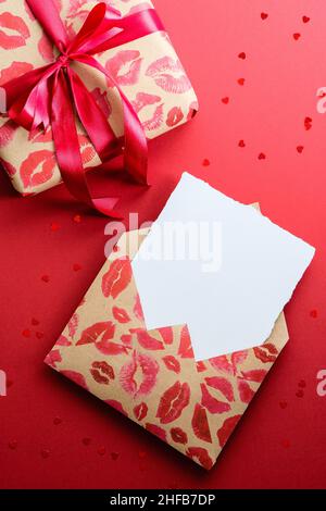 Happy Valentines Day Concept.Craft Papierumschlag mit Lippenstiftmarkierungen mit leeren Papierkarte innen, Geschenkbox gebunden roten Band auf rotem Hintergrund. Flach l Stockfoto