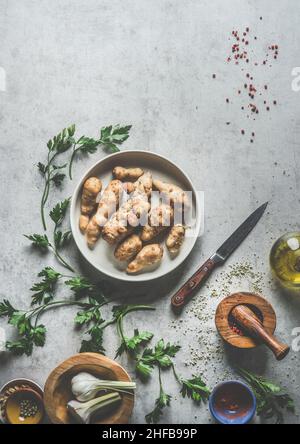 Jerusalemer Artischocke in Schüssel auf grauem Beton Küchentisch mit Zutaten, Petersilie, Mörser und Stößel, Küchenmesser, Öl und Knoblauch. Vege wird vorbereitet Stockfoto