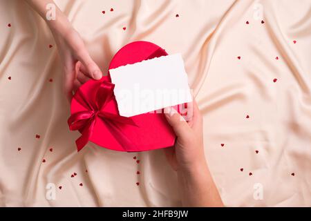 Der Mann gibt einer Frau eine herzförmige Schachtel mit einem leeren Grußkartenmockup. Alles gute zum Valentinstag, Muttertag, Geburtstagskonzept Stockfoto