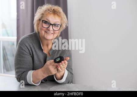 Lächelnde ältere Frau, die zu Hause das Pulsoximeter verwendet, die Sauerstoffsättigung selbst misst, die Pulsfrequenz überprüft und während der Messung Gesundheitsindikatoren kontrolliert Stockfoto