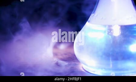 Nahaufnahme eines durchsichtigen Glasflaschens mit Shisha auf dem Tisch mit einer Rauchwolke. Shisha-Flasche unter dem blauen Lampenlicht, das auf einem Holztisch steht. Stockfoto