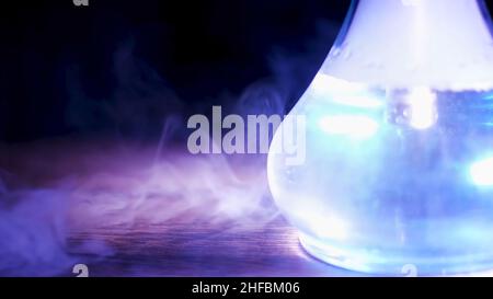 Nahaufnahme eines durchsichtigen Glasflaschens mit Shisha auf dem Tisch mit einer Rauchwolke. Shisha-Flasche unter dem blauen Lampenlicht, das auf einem Holztisch steht. Stockfoto