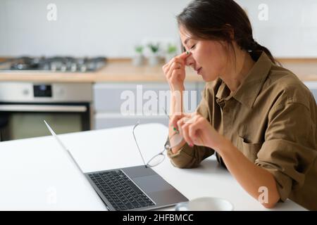Eine asiatische Frau oder Studentin schloss die Augen und massierte ihre Nase, wobei sie an schweren Kopfschmerzen, Migräne, litt. Müde junge Frau mit Computerarbeit überwältigt Stockfoto