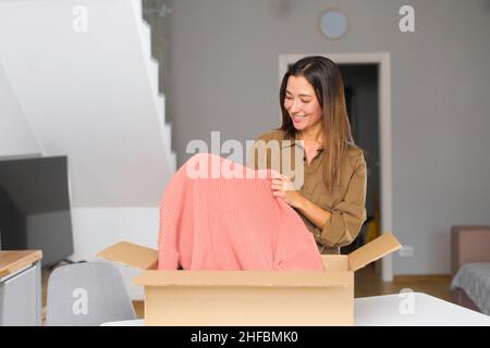 Positive junge asiatische Frau, die mit einem Karton steht und neue Kleidung auszieht, während sie neugierig auf bestellte Artikel aus einem Online-Shop ist. Lächelnder Kunde zufrieden mit schnellem Kurierdienst Stockfoto