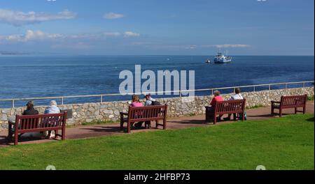 Menschen genießen den Meerblick von Shaldon, South Devon, während ein Frachtschiff vom Hafen von Teignmouth abfährt. Stockfoto