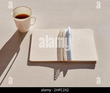 Öffnen Sie ein leeres Notizbuch und eine kleine weiße Tasse mit Kaffee auf einem weißen Tisch bei hellem Sonnenlicht Stockfoto