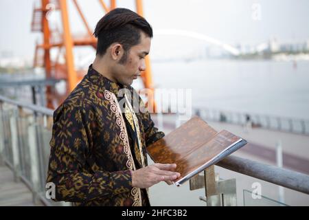 Ein junges männliches Model in indonesischer Batik mit balinesischem Blumenmuster liest ein Buch über Batik im Long Museum, West Bund, Shanghai China Stockfoto