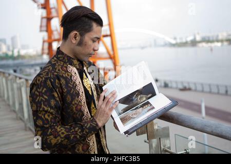 Ein junges männliches Model in indonesischer Batik mit balinesischem Blumenmuster liest ein Buch über Batik im Long Museum, West Bund, Shanghai China Stockfoto