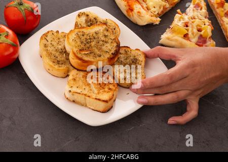 Hand nehmen ein knuspriges Knoblauchbrot mit Orangano, im Hintergrund Scheiben Pizza mit Käse, Ananas und Schinken, Dekoration mit Tomaten, Tisch Stockfoto