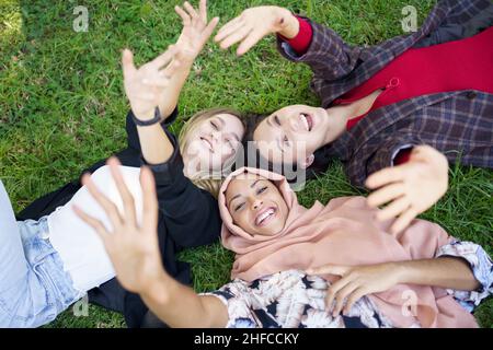 Fröhliche junge, vielfältige Freundinnen, die auf Gras liegen und die Kamera anlächeln Stockfoto