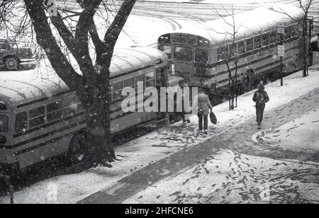 1970s, historisch, winterlich und inmitten von eiskalten Schnee, warten draußen auf der Straße zwei Schulbusse für Schüler, Harlan, Iowa, USA. Die Bereitstellung von Transport für Schulkinder ist in Amerika ein riesiges Unterfangen. Ungewöhnlich in einem kapitalistisch angetriebenen Land war es traditionell ein sozialisiertes System, bei dem lokale öffentliche Einrichtungen kostenlose Schultransporte für Kinder anbieten - manchmal abhängig davon, wie weit sie von den Schulen leben - aus lokalen Steuergeldern und Bundesmitteln. Der Schulbus ist ein ikonischer Teil der amerikanischen Kultur Stockfoto