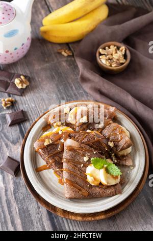 Wir feiern den Pfannkuchen-Tag, ein gesundes Frühstück. Köstliche hausgemachte Schokoladencrepes mit Nüssen, Karamell, Bananen auf rustikalem Holztisch. Stockfoto