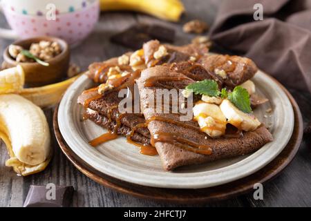 Wir feiern den Pfannkuchen-Tag, ein gesundes Frühstück. Köstliche hausgemachte Schokoladencrepes mit Nüssen, Karamell, Bananen auf rustikalem Holztisch. Stockfoto