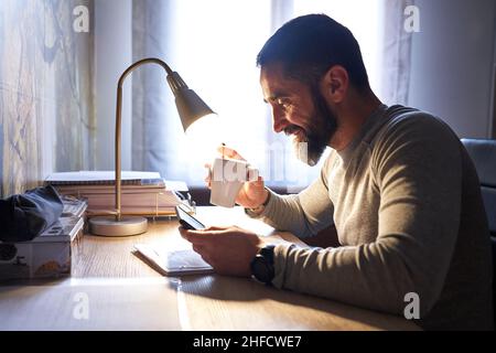 Junger weißer Mann mit einem ordentlichen Bart, der während einer Infusion am Schreibtisch studiert und das Telefon beobachtet Stockfoto