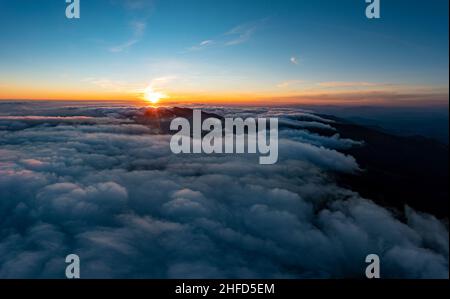 Sonnenlicht, das hell auf dem blauen Himmel über einer dicken Schicht weißer, flauschiger Wolken bei Sonnenaufgang leuchtet, mit Bergen im Hintergrund Stockfoto