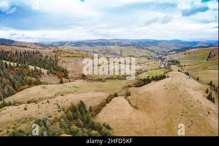 Hügel Hänge mit grünen vergilbten Wäldern bedeckt Felder Wiesen und entwaldeten Gebieten rund um Dorf im Tal unter bewölktem Herbsthimmel Stockfoto