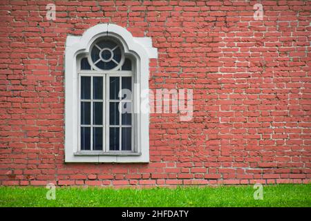 Backsteinmauer mit nur einem Fenster zur Hausfront. Fassade eines alten Gebäudes und einer grünen Wiese. Das Äußere des alten Hauses. Stockfoto