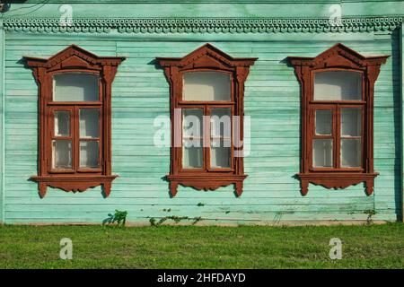 Drei Fenster in einer Reihe an der Wand eines alten russischen Hauses, das mit Holzschnitzereien verziert und in aqua- und braunen Farben gestrichen ist Stockfoto