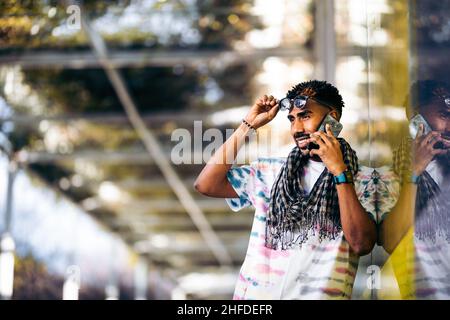 Lächelnder schwarzer Mann, der sich mit seinem Smartphone an eine Glaswand lehnt. Seine Brille mit der Hand ausziehen. Stockfoto