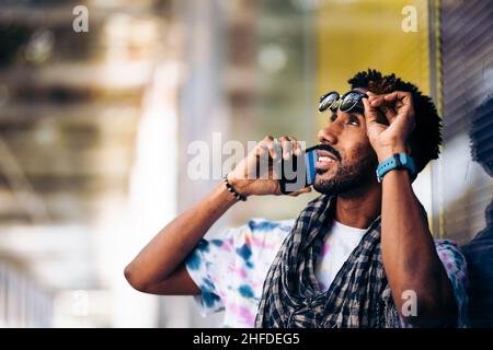 Nahaufnahme eines lächelnden schwarzen Mannes, der sich mit seinem Smartphone im Profil an einer Glaswand lehnt und seine Brille auszieht Stockfoto