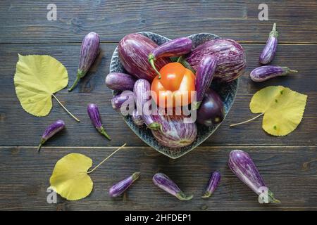 Herbstgemüse. Roter Paprika, verschiedene Auberginen-Gemüsesorten in herzförmiger Platte und gelbe Blätter auf dunklem rustikalem Tisch. Gesund, V Stockfoto