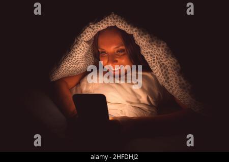 Glückliche lächelnde Frau, die im Bett unter der Decke lag und nachts vor dem Schlafen auf dem Smartphone tippte. Sucht nach sozialen Medien und Kommunikation Stockfoto