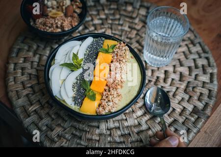 Vogelansicht einer grünen veganen Smoothie-Schale mit tropischen Früchten, Müsli, Mango, Spirulina, Kokosnuss, Nüssen, chia-Samen, Minze auf einem hölzernen Frühstückstisch Stockfoto