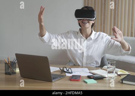 Fröhliche junge Geschäftsfrau, die die Hände in der Luft bewegt, während sie am Arbeitsplatz eine Virtual-Reality-Brille verwendet Stockfoto