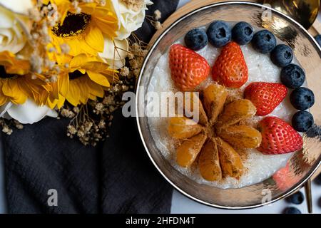 Weiße Smoothie-Schüssel mit Reis, Zimt, Orange, Heidelbeeren und Erdbeeren auf der Oberseite und gelben Blüten Stockfoto