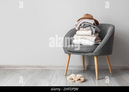 Stapel von warmen Pullovern mit Hut auf Sessel und Pantoffeln in der Nähe der hellen Wand Stockfoto