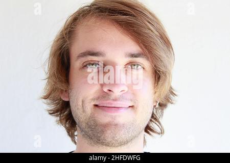 Junger Mann mit grünen Augen, der offen lächelt. Junger Mann mit langen blonden Haaren lächelt offen isoliert auf weißem Hintergrund. Gesichtsausdrücke. Stockfoto