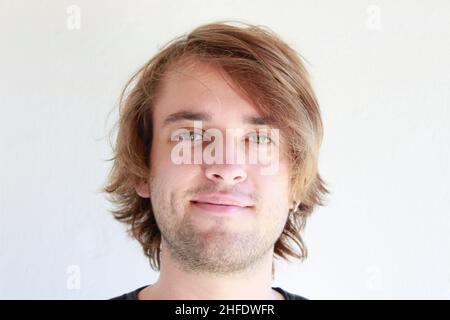 Junger Mann mit grünen Augen, der offen lächelt. Junger Mann mit langen blonden Haaren lächelt offen isoliert auf weißem Hintergrund. Gesichtsausdrücke. Stockfoto