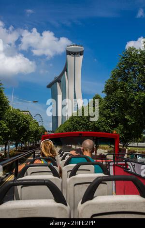 Besucher im offenen Bus besichtigen die berühmten Marina Bay Sands und eine Reihe üppig grüner Bäume entlang der Straße, Singapur. Stockfoto