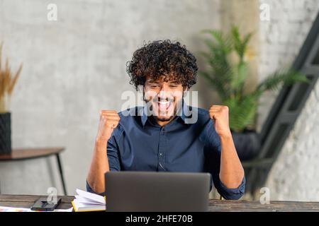 Aufgeregter pakistanischer Geschäftsmann, der auf den Laptop-Bildschirm schaut und sich freut. Glücklicher Kerl schreiend ja starrend mit geöffnetem Mund auf Monitor amüsiert mit unerwarteter Gelegenheit Stockfoto