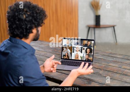 Remote-Meeting per Videoanruf. Indischer Mann, der in der Küche sitzt und die App auf dem Laptop für die Online-Kommunikation mit einer Gruppe verschiedener multirassischer Menschen nutzt. Rückansicht Stockfoto