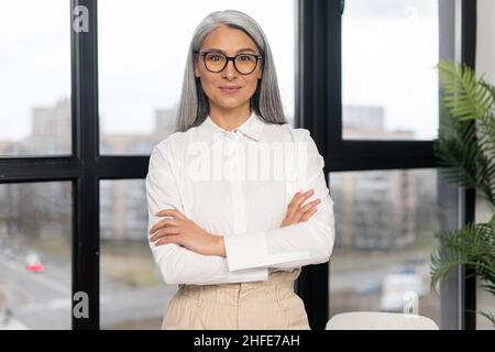 Seriöse und strenge alte Geschäftsfrau mit elegantem Casual-Hemd und stilvoller Brille, die mit gekreuzten Armen im modernen Bürobereich steht. Reife herrische Dame, ältere zielstrebige Führerin Konzept Stockfoto