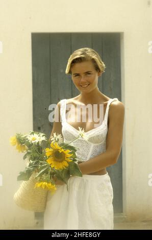 Attraktives blondes Haar junge Frau Porträt lächelnde vordere Kamera trägt weißes Kleid tiefer Ausschnitt mit Sonnenblumenstrohkorb Frühlingsferien Stockfoto