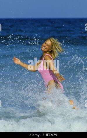 Dynamische blonde Haare junge Frau Schönheit lächelnd Profil Front-Kamera läuft spielen spritzt Wellen blau Meerwasser Hintergrund Stockfoto